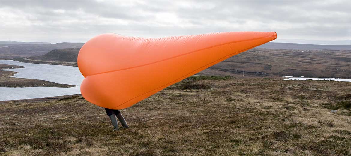 Link to Into The Wind project by Michael Cross. Image shows a man holding a large fabric funnel which is filled by a strong wind, he is leaning over into the wind.